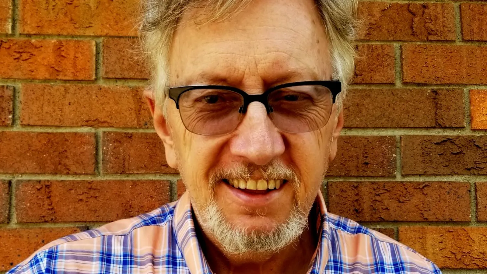 John Low wearing glasses, with brick background and plaid shirt