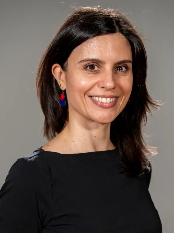 Portrait of a woman with dark shoulder-length hair wearing a black top and red-and-blue earrings, smiling against a gray background.